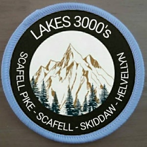 May include: A blue and black round patch with the text "LAKES 3000'S" in white. The patch features a white mountain range with brown peaks and green trees against a white background. The text "SCAFELL PIKE - SCAFELL - SKIDDAW - HELLVELLYN" is written in a circle around the mountain range.