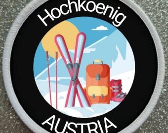 May include: A white and black circular patch with the text "Hochkoenig Austria" in white. The patch features a cartoon illustration of a snowy mountain scene with skis, ski poles, a backpack, and ski boots.