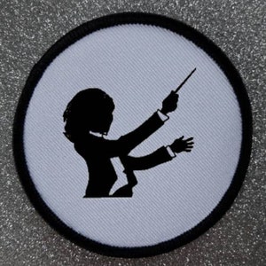Könnte beinhalten: Ein weißer runder Patch mit schwarzem Rand. Der Patch zeigt eine schwarze Silhouette eines Dirigenten, der ein Orchester leitet.