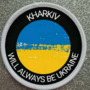 Puede incluir: Parche circular blanco con borde negro y el texto "KHARKIV WILL ALWAYS BE UKRAINE" en blanco. El centro presenta la bandera ucraniana, azul y amarilla.