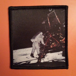 Op de afbeelding: Een zwart-witfoto van een astronaut die op de maan loopt. De astronaut draagt een wit ruimtepak en loopt naar een maanlander. De foto is gedrukt op een zwarte stoffen patch met een zwarte rand.
