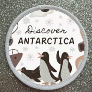 May include: A white round patch with a grey border. The patch has a cartoon illustration of penguins and snowflakes. The text "Discover Antarctica" is printed in black.