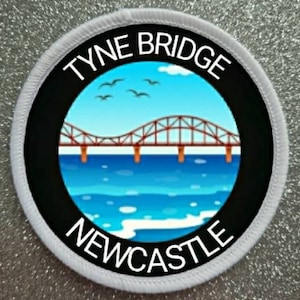 Puede incluir: Un parche circular blanco con texto negro y un círculo negro en el centro. El texto dice "TYNE BRIDGE NEWCASTLE". El círculo negro contiene una imagen de dibujos animados de un puente rojo sobre una masa de agua azul con olas blancas.