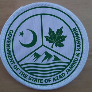Op de afbeelding: Een witte ronde patch met groene tekst en symbolen. De tekst luidt "Government of the State of Azad Jammu & Kashmir". De symbolen omvatten een halve maan met een ster, een esdoornblad en drie bergen met golven eronder.