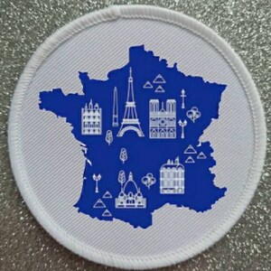 Könnte beinhalten: Ein weißer gestickter Aufnäher mit einer blauen Umrandung von Frankreich und weißen Symbolen berühmter Sehenswürdigkeiten, darunter der Eiffelturm, die Kathedrale Notre-Dame und der Arc de Triomphe.