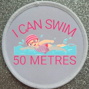 May include: A round, gray patch with a white border. The patch features a cartoon swimmer in a pink swimsuit and cap, swimming in blue water. The text "I CAN SWIM 50 METRES" is in pink.