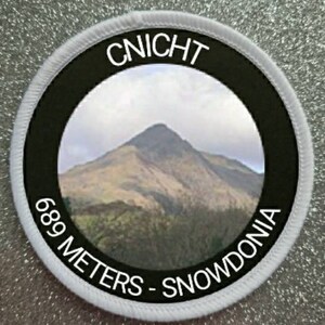 Könnte beinhalten: Ein runder weißer und schwarzer Aufnäher mit dem Text "CNICHT 689 METERS - SNOWDONIA" und einem Foto eines Berges in der Mitte.