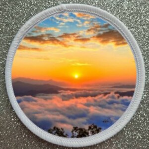 Może przedstawiać: Biała okrągła łatka z kolorowym wschodem słońca nad morzem chmur. Niebo jest w kolorze niebieskim, pomarańczowym i żółtym. Słońce jest jasnym pomarańczowym okręgiem w środku obrazu.