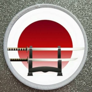 May include: A white round patch with a red and white Japanese rising sun design. Two silver swords are displayed on a black stand in front of the sun.