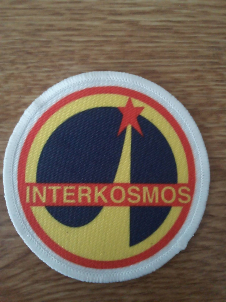 K&ouml;nnte beinhalten: Ein wei&szlig;er, roter und gelber runder Patch mit dem Text "INTERKOSMOS" in einem schwarzen Kreis. Ein roter Stern mit gelber Umrandung befindet sich in der oberen rechten Ecke des Kreises.