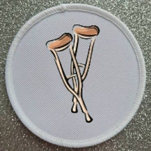 Puede incluir: Un parche redondo blanco con una ilustración marrón y negra de un par de muletas.