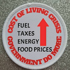 May include: A circular patch with white edging and a grey center. Red text encircles the edge, reading "COST OF LIVING CRISIS" and "GOVERNMENT DO MORE". The center lists "FUEL TAXES ENERGY FOOD PRICES" with a red arrow.