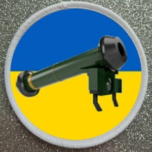 May include: A white circular patch with a blue and yellow Ukrainian flag design. A green and black cartoon rocket launcher is positioned on top of the flag.