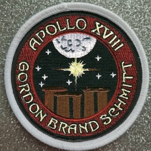 May include: An embroidered patch in red, white and black with the text "Apollo XVIII Gordon Brand Schmitt" and a depiction of the moon, stars and three pillars.