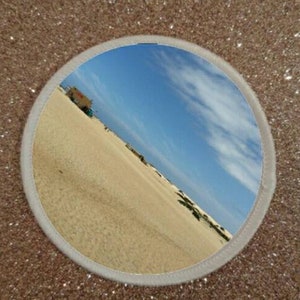 May include: A round, white-framed mirror reflecting a blue sky with white clouds and a sandy beach with a small building in the distance.
