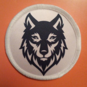 Peut inclure: Un écusson circulaire blanc avec un motif de tête de loup noir et blanc. La tête du loup est tournée vers l'avant, la gueule fermée. L'écusson a une bordure blanche.