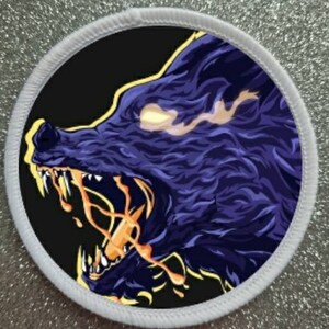 May include: Round patch featuring a stylized wolf's head in profile. The wolf is rendered in shades of purple and black, with glowing yellow eyes and fiery orange details emanating from its mouth. The patch has a white border.