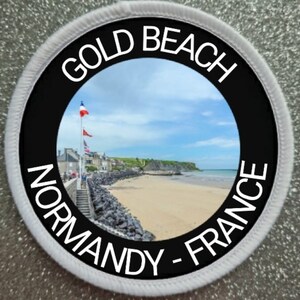 May include: A white and black circular patch with the text "Gold Beach Normandy - France" around the edge. The centre of the patch features a photo of a beach with a town in the background.