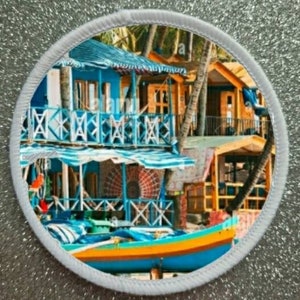 May include: A colorful round coaster with a beach scene featuring a blue and yellow boat, palm trees, and a blue and yellow building with a wooden balcony.