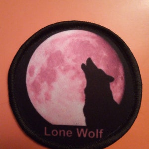 May include: Black and white embroidered patch featuring a wolf howling at a pink full moon. The patch has a black border and the words "Lone Wolf" are embroidered in white below the moon.