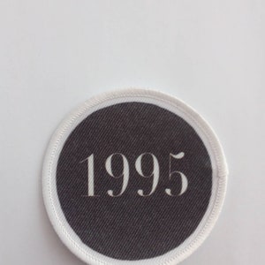 Op de afbeelding: Een witte ronde patch met een zwarte achtergrond en het getal 1995 in wit gedrukt.