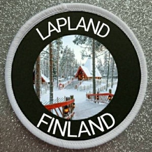 May include: Circular patch with a black centre and white border. The patch displays the words "LAPLAND" and "FINLAND" in white, surrounding a winter scene with snow-covered trees and buildings.