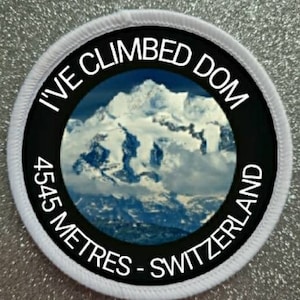 Könnte beinhalten: Runder weißer Aufnäher mit schwarzem Text "I've Climbed Dom 4545 Metres - Switzerland" und einem Foto eines schneebedeckten Berggipfels.