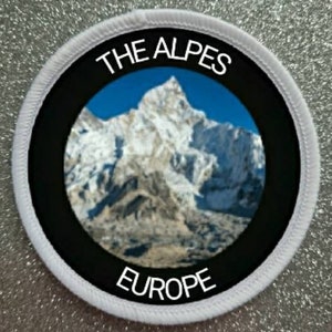 May include: White round patch with black border and white text. The text reads "THE ALPES EUROPE". The center of the patch features a photo of a snow-capped mountain range.