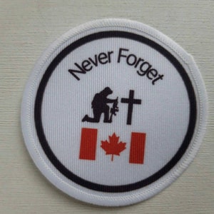 Può includere: Toppa rotonda bianca con bordo nero. La toppa presenta la scritta "Never Forget" sopra una sagoma di soldato inginocchiato, una croce e la bandiera canadese. La toppa è probabilmente un oggetto commemorativo.