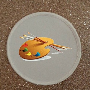 Op de afbeelding: Een ronde, witte patch met een bruin en geel palet en twee kwasten. Het palet heeft kleurrijke verfspatten.