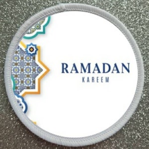Puede incluir: Parche redondo blanco con un patrón geométrico azul y amarillo en el lado izquierdo y el texto "RAMADAN KAREEM" en letras azules en el lado derecho.
