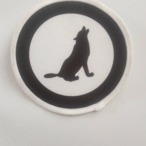 Könnte beinhalten: Ein weißer runder Patch mit schwarzem Rand und einer schwarzen Silhouette eines Wolfes, der zum Mond heult.
