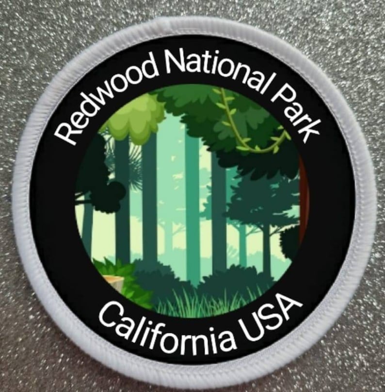Pu&ograve; includere: Un cerotto rotondo bianco con testo nero e un cerchio nero al centro. Il testo recita "Redwood National Park California USA". Il cerchio nero contiene un'illustrazione verde e blu di una foresta.