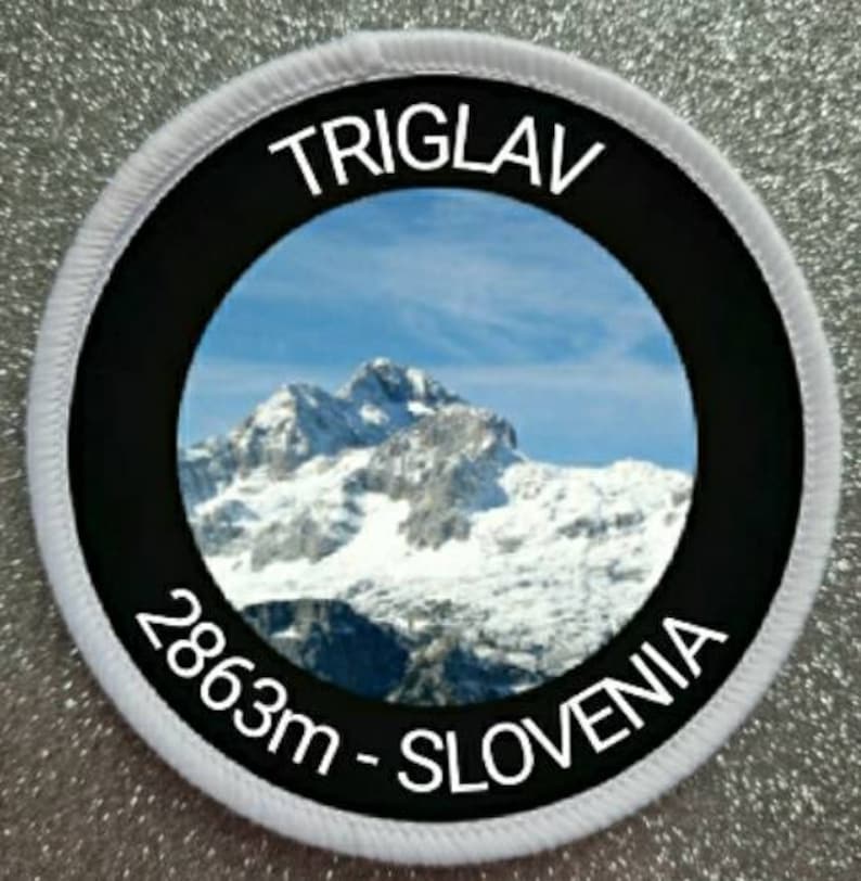 以下が含まれることがあります： 上部に「TRIGLAV」、下部に「2863m - SLOVENIA」と書かれた丸いパッチ。中央には、青い空の下、雪をかぶった山々が描かれています。パッチは黒い中央リングと白い縁取りが施されています。