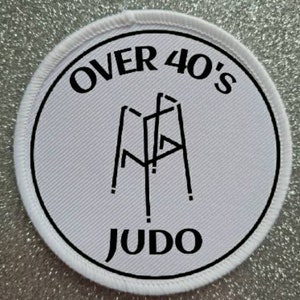 Puede incluir: Parche redondo blanco con texto negro y un símbolo de judo. El texto dice "Over 40's Judo".