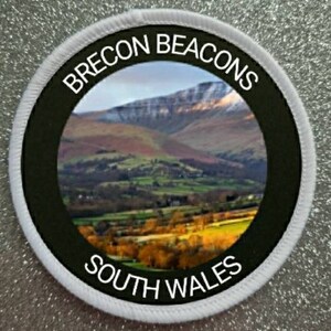 Op de afbeelding: Een witte ronde patch met zwarte tekst en een zwarte rand. De tekst luidt "Brecon Beacons South Wales". Het midden van de patch toont een foto van een bergketen met een vallei op de voorgrond.