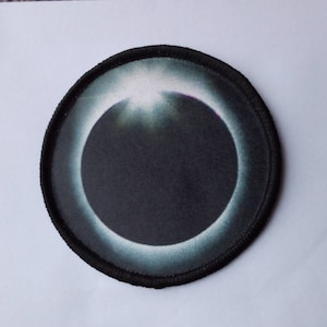 Può includere: Un cerotto circolare bianco e nero con una grafica di un'eclissi solare. L'eclissi è rappresentata da un cerchio scuro circondato da un anello bianco.