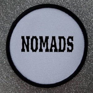 May include: White round patch with black stitching and the word "NOMADS" in black block letters.