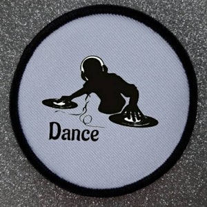 Könnte beinhalten: Runder weißer Patch mit schwarzem Rand, der die Silhouette eines DJs mit Kopfhörern und Plattenspielern zeigt. Der Text "Dance" ist unter dem DJ gedruckt.
