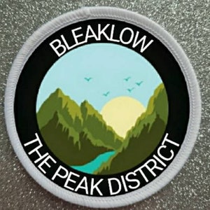 Op de afbeelding: Ronde witte patch met zwarte tekst en een groene en blauwe berg scène. De tekst luidt "Bleaklow The Peak District".