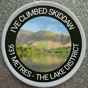 Può includere: Una toppa rotonda bianca e nera con il testo "I've Climbed Skiddaw 937 Metres - The Lake District". La toppa presenta una foto di una catena montuosa e un lago.