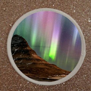 Può includere: Una toppa rotonda con un bordo bianco che mostra un'aurora boreale vibrante in tonalità di verde, viola e rosa sopra una catena montuosa scura.