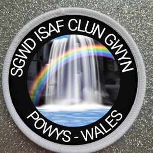 May include: A white circular patch with black text and a black circle in the centre. The text reads "SGWD ISAF CLUN GWYN POWYS - WALES". The black circle contains a photo of a waterfall with a rainbow.