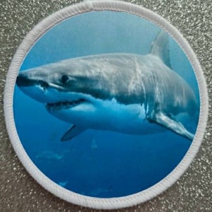 May include: A white great white shark swims in blue water. The shark is facing the left side of the image and has its mouth open.