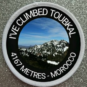 May include: White round patch with black text and a photo of a mountain range. The text reads "I've Climbed Toubkal 4167 Metres - Morocco".