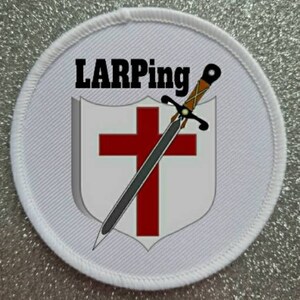 May include: White round patch with a red cross and a sword on a white shield. The text "LARPing" is at the top of the patch.