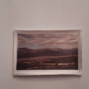May include: A white fabric patch with a stitched border, featuring a printed image of a landscape with a lake, hills, and a cloudy sky.