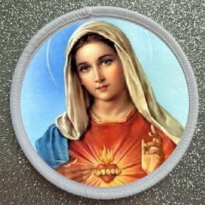May include: A white and blue religious patch with a depiction of the Virgin Mary with a flaming heart. The heart is surrounded by a golden halo and the Virgin Mary is wearing a blue robe and a white veil.