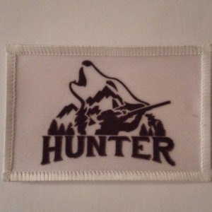 May include: A rectangular white patch with a black graphic of a howling wolf over a mountain range. The word "HUNTER" is printed in bold black letters below the graphic. The patch has a white stitched border.