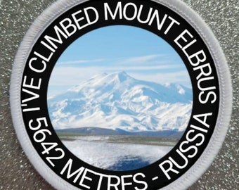 Peut inclure: Un écusson rond avec une bordure blanche et un centre noir. L'écusson présente un paysage de montagne enneigée et le texte "I'VE CLIMBED MOUNT ELBRUS RUSSIA 5642 METRES". L'image montre une chaîne de montagnes sous un ciel bleu.